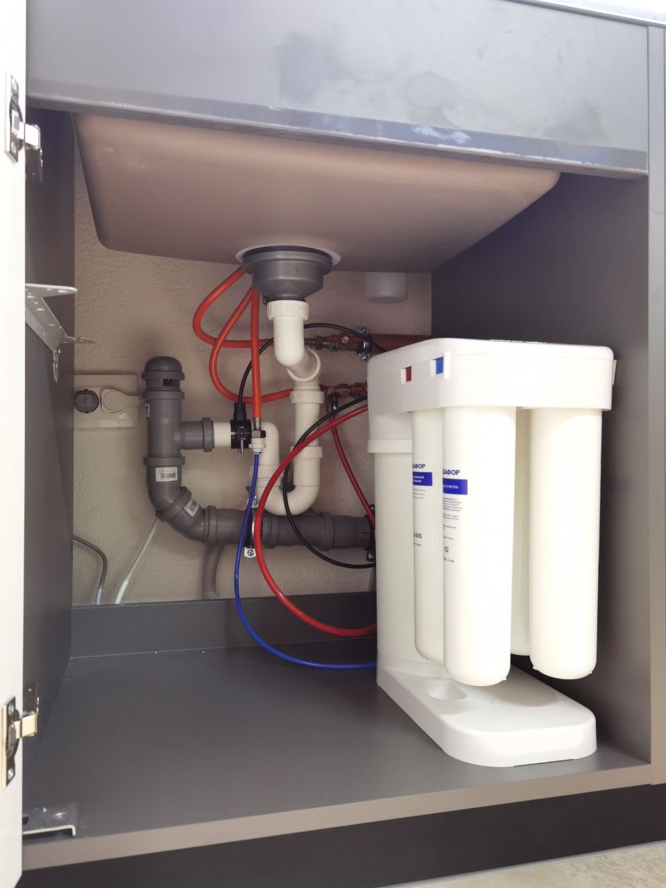 Filtr wody pod zlewem, widoczne rury doprowadzające wodę, szary syfon i biały system filtracji z trzema cylindrami w szafce pod zlewem.