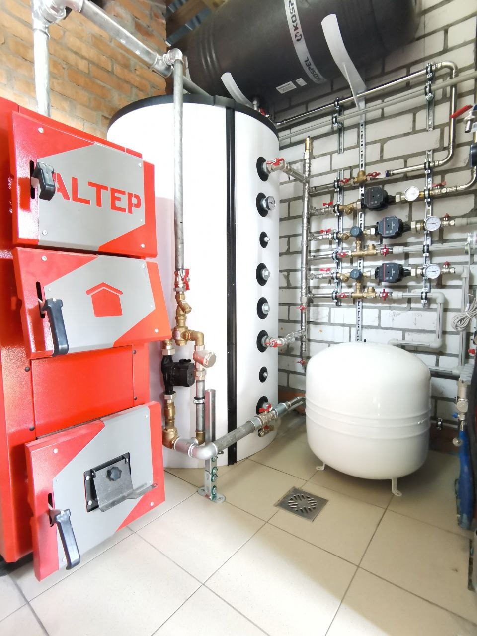 Kotłownia z piecem Altek, zasobnikiem ciepłej wody Kospel i rozbudowaną instalacją. Widoczne liczniki ciepła, rury miedziane i biała cegła na ścianie.