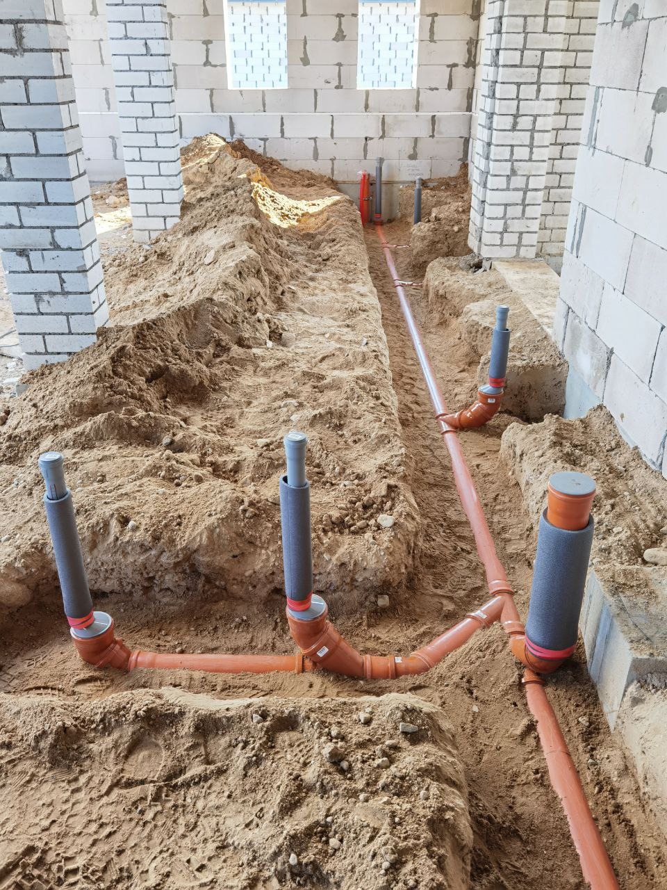 Instalacja kanalizacyjna w wykopie ziemnym, pomarańczowe rury z szarymi izolacjami pionowymi, surowe ściany z białego bloczka w tle, widok z góry.