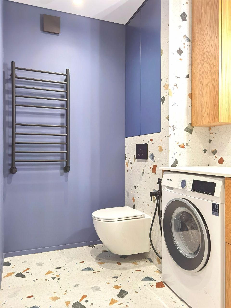 Nowoczesna łazienka w odcieniach błękitu i terazzo: wisząca toaleta, grzejnik drabinkowy, pralka i szafki. Uwagę zwraca wzór terazzo na podłodze i ścianie.