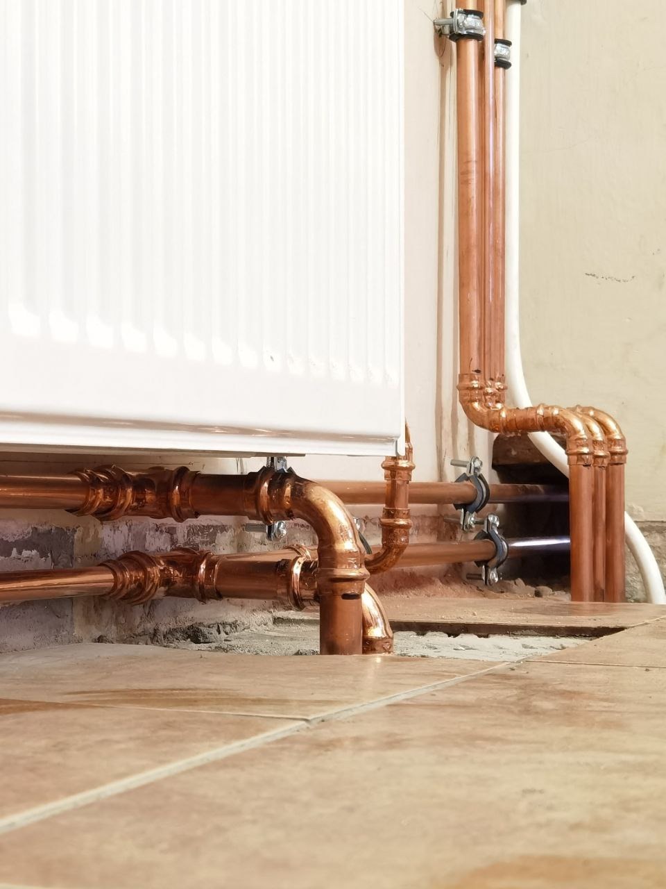 Miedziane rury grzewcze przy białym grzejniku, zamocowane do ściany klamrami, widoczne na tle beżowych płytek podłogowych i fragmentu ściany.