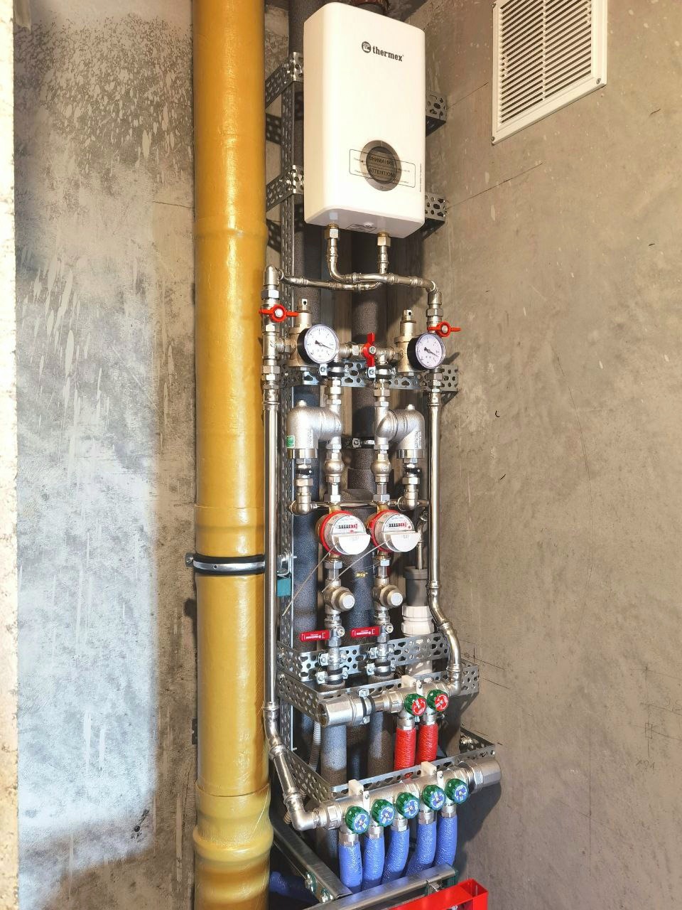 Kompleksowa instalacja hydrauliczna z licznymi zaworami, manometrami i filtrami, zamontowana na ścianie o surowym wykończeniu. Widoczny bojler marki Thermex.