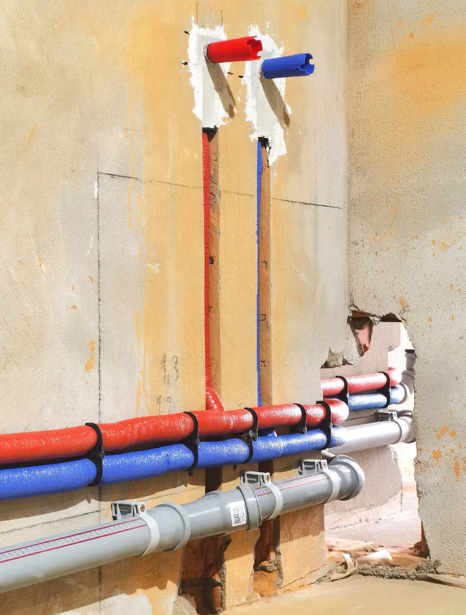 Instalacja wodna: rury czerwone i niebieskie prowadzone w ścianie, zabezpieczone uchwytami. Szare rury kanalizacyjne na dole. Surowy tynk w tle.