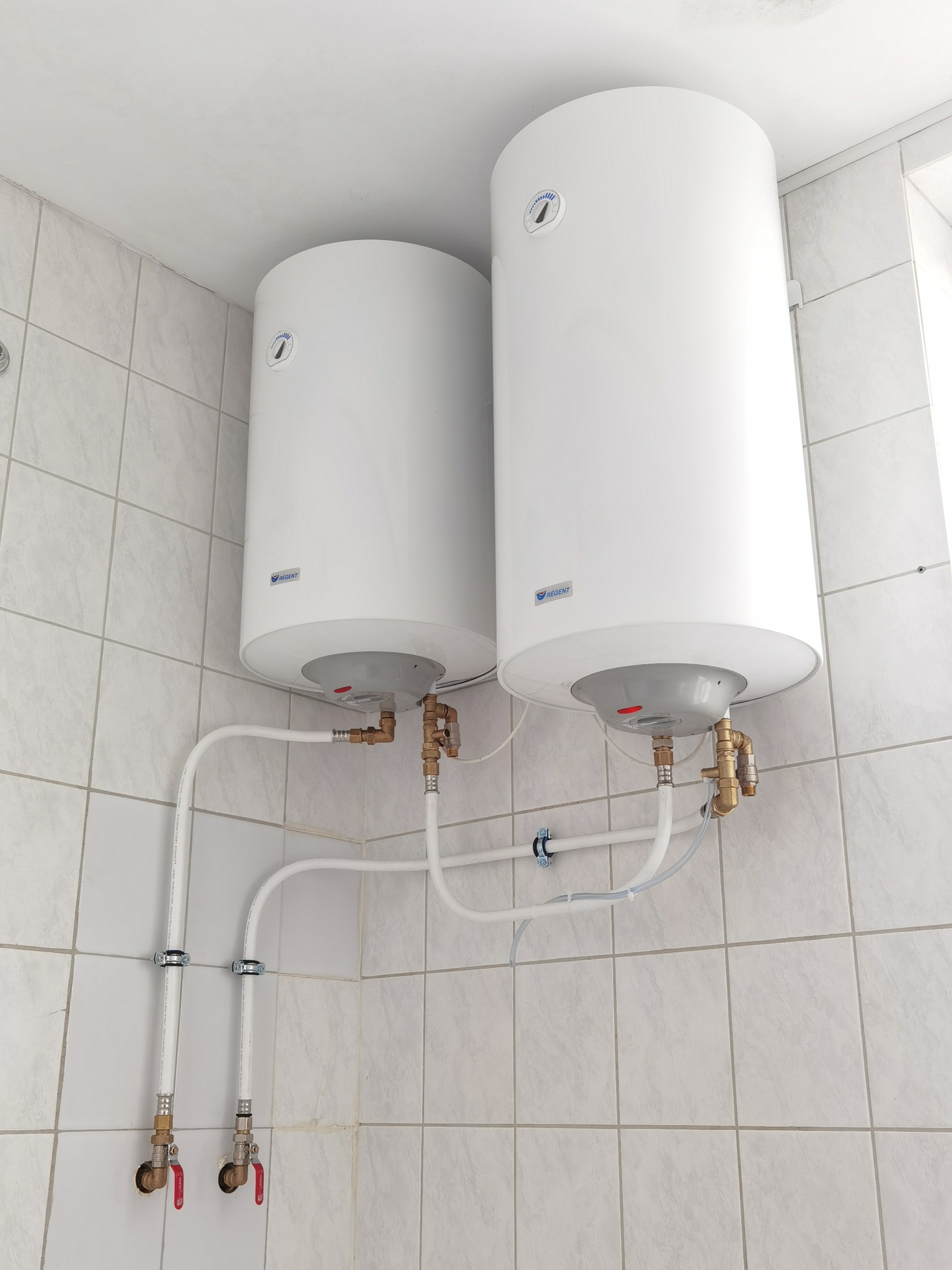 Dwa białe, cylindryczne bojlery elektryczne zamontowane w rogu pomieszczenia z jasnymi, kwadratowymi płytkami. Widoczne rury i zawory doprowadzające wodę.