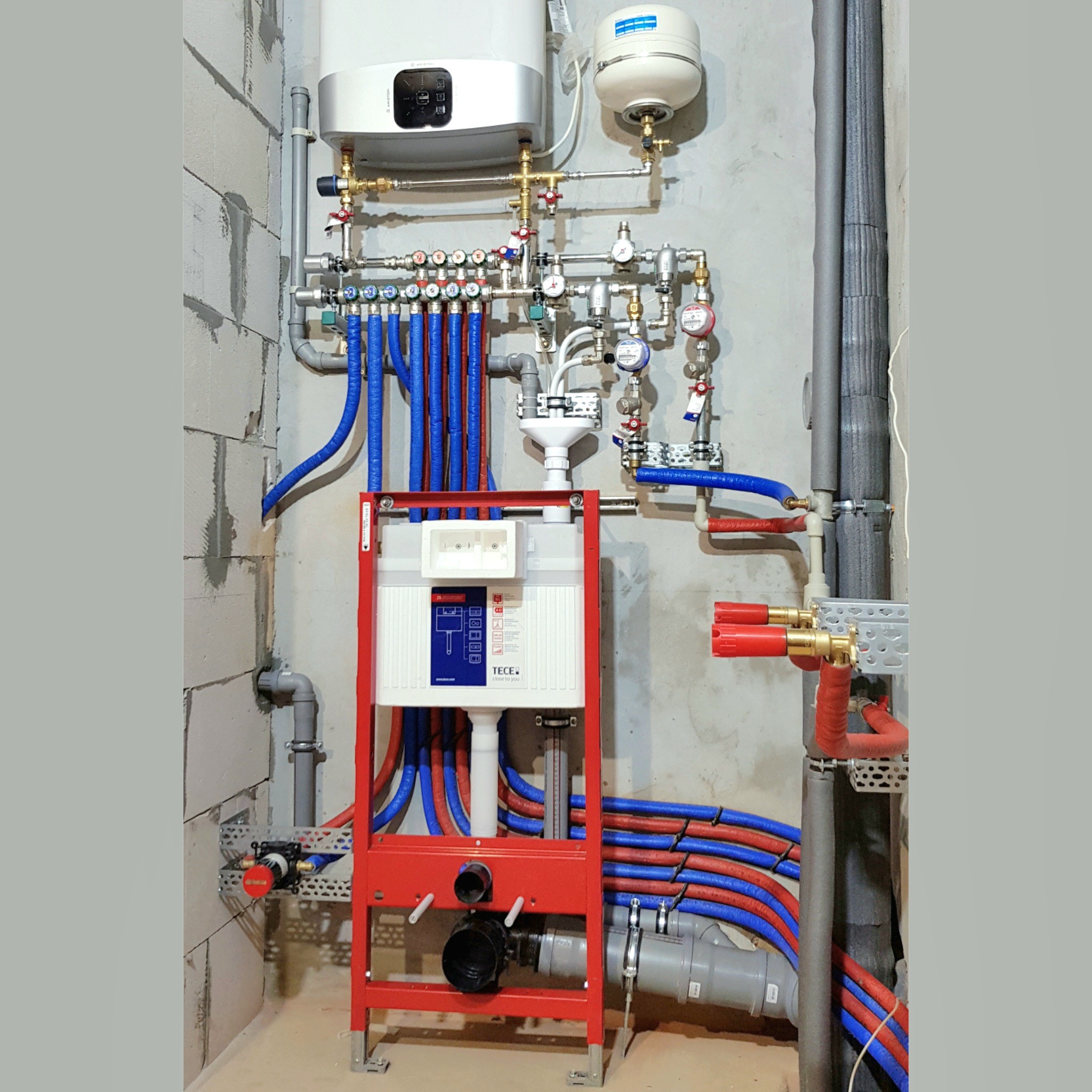 Kompleksowa instalacja wodno-kanalizacyjna z czerwonym stelażem podtynkowym TECE, rozdzielaczami i bojlerem Ariston na tle ściany z betonu komórkowego.