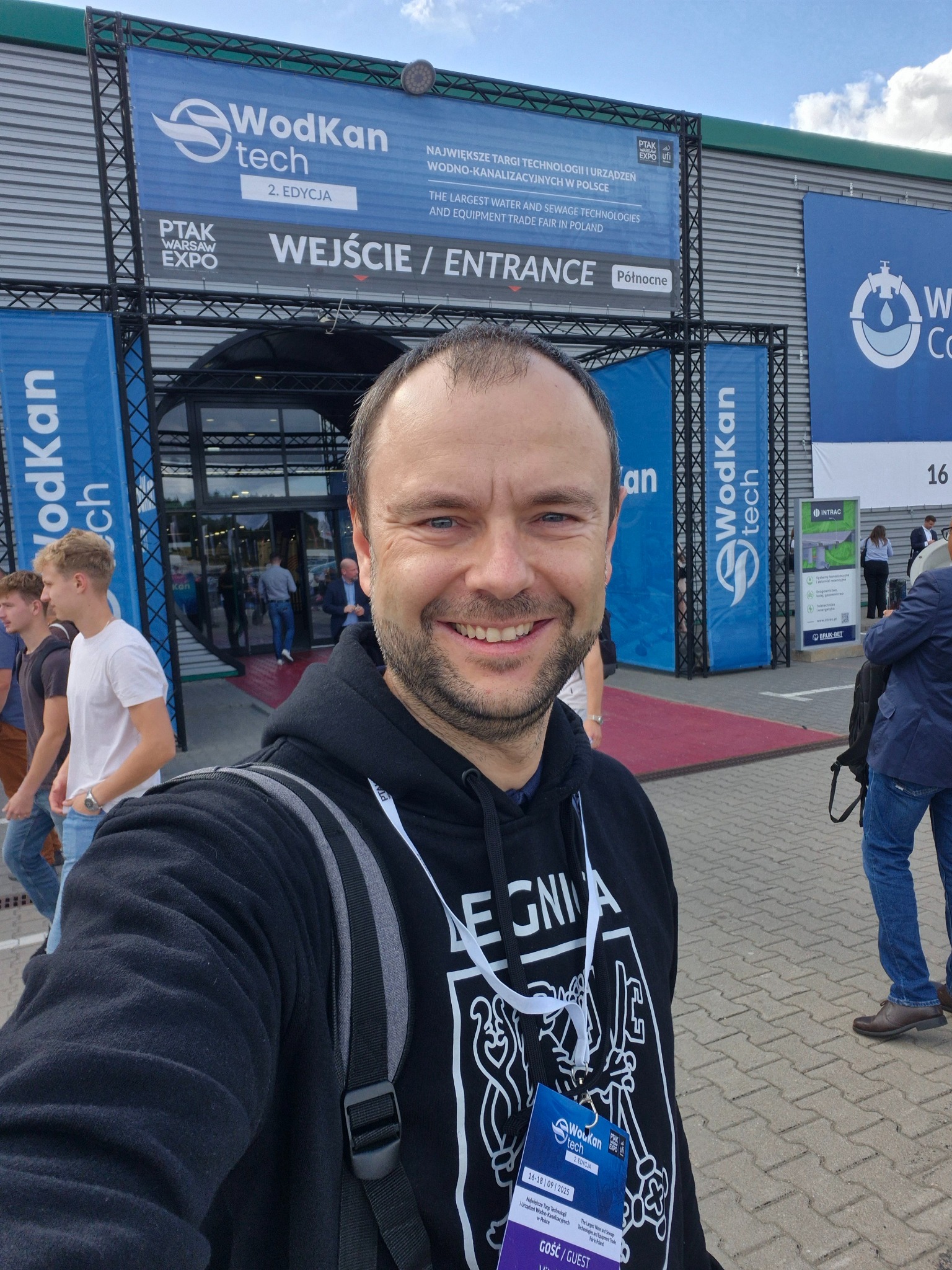 Uśmiechnięty mężczyzna na tle wejścia na targi Wod-Kan Tech, z widocznymi banerami i logotypami. Widoczny tłum ludzi w tle, pochmurne niebo.