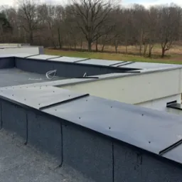 Wykończony dach płaski z papy termozgrzewalnej i obróbek blacharskich, widok z góry na tle drzew i niebieskiego namiotu. Detal dachu i kominów wentylacyjnych.