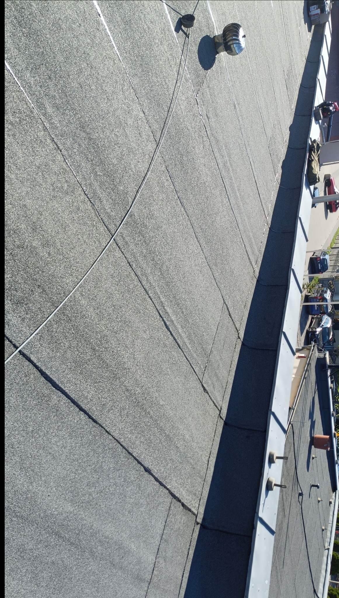 Szary, płaski dach pokryty papą termozgrzewalną z widocznymi kominkami wentylacyjnymi i elementami obróbek blacharskich, sfotografowany z góry pod kątem.