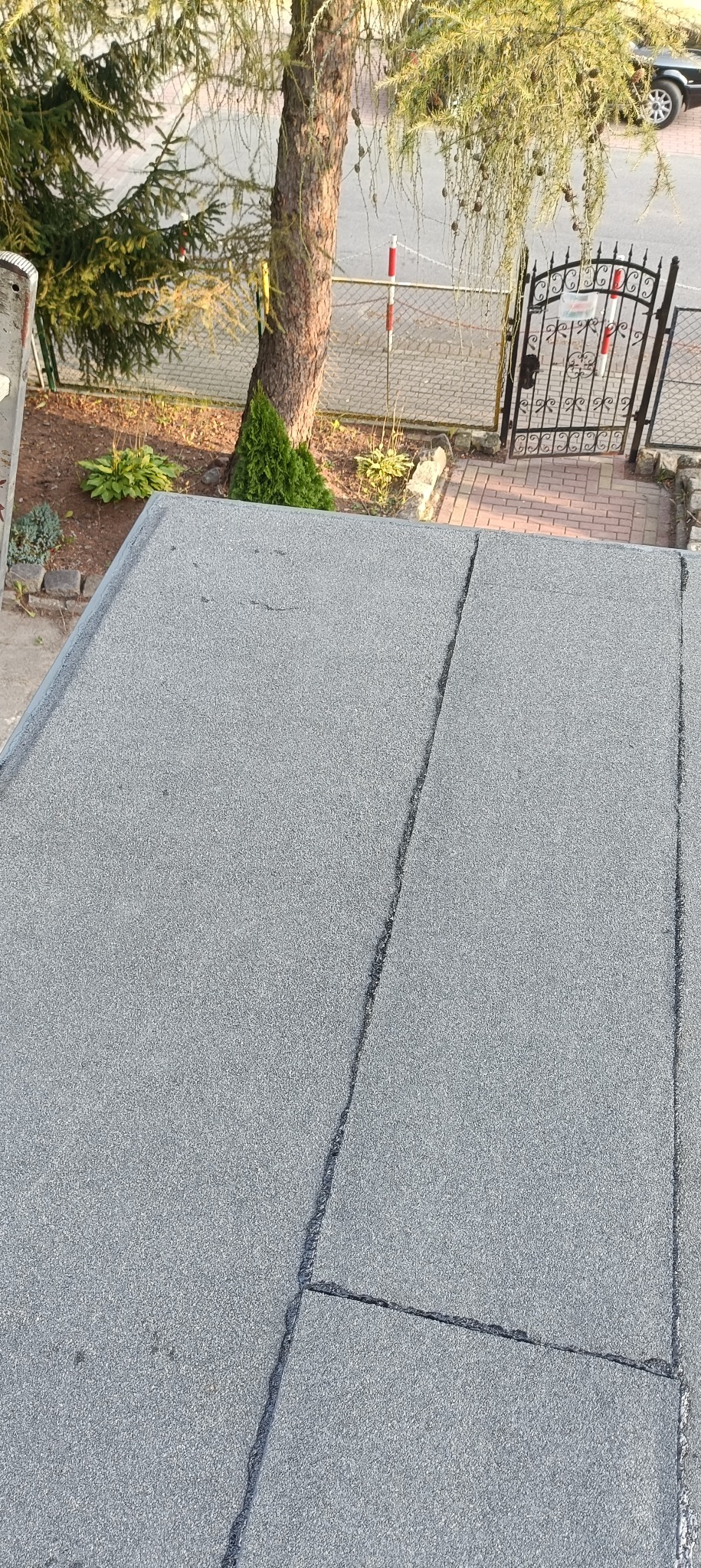 Szary, płaski dach z papy widziany z góry, z widocznymi łączeniami, na tle drzewa i ogrodzenia. Specyfika ujęcia podkreśla fakturę dachu.