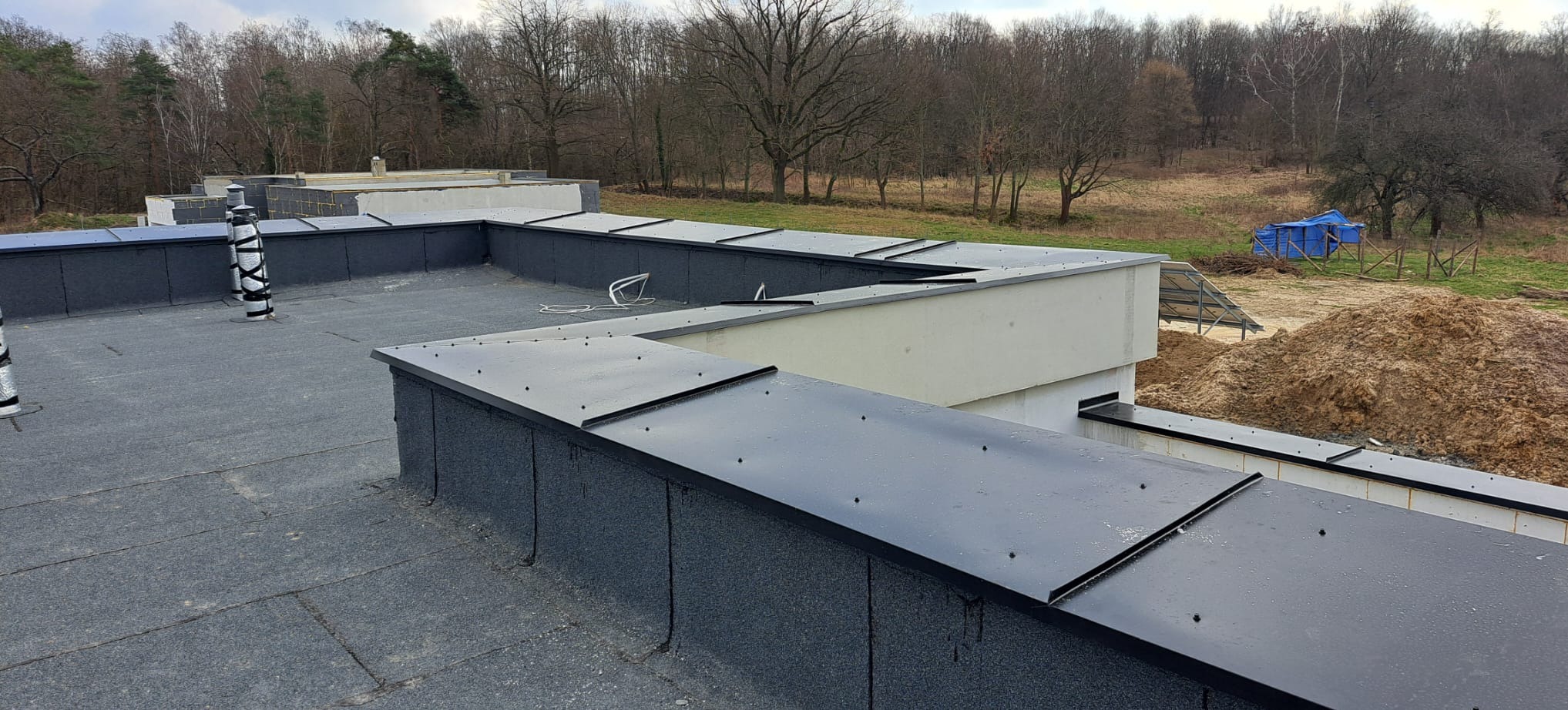 Wykończony dach płaski z papy termozgrzewalnej i obróbek blacharskich, widok z góry na tle drzew i niebieskiego namiotu. Detal dachu i kominów wentylacyjnych.