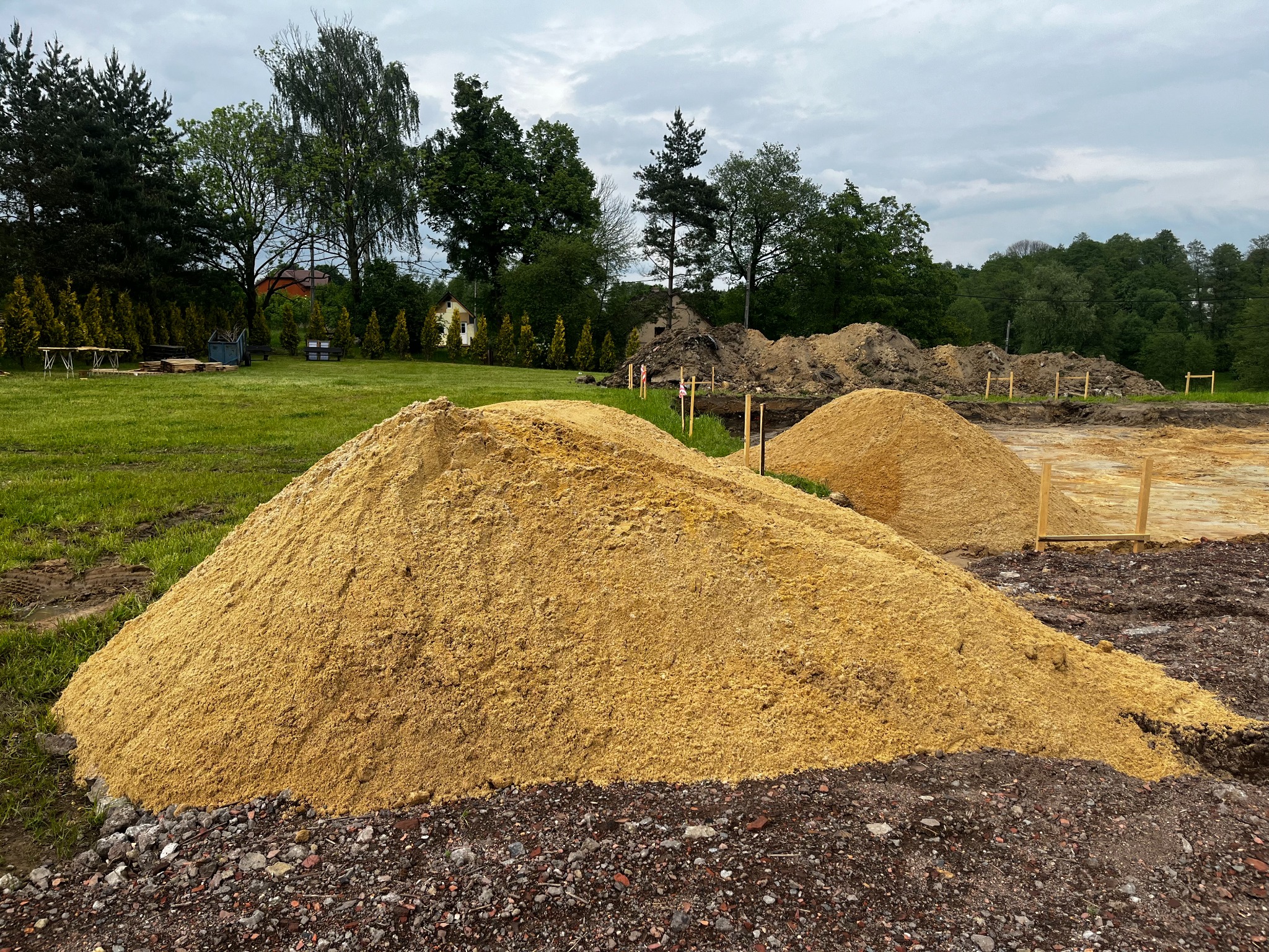 Dwa kopce przesiewanego materiału budowlanego na placu budowy, widoczne drewniane ramy fundamentów i zieleń w tle pod pochmurnym niebem.