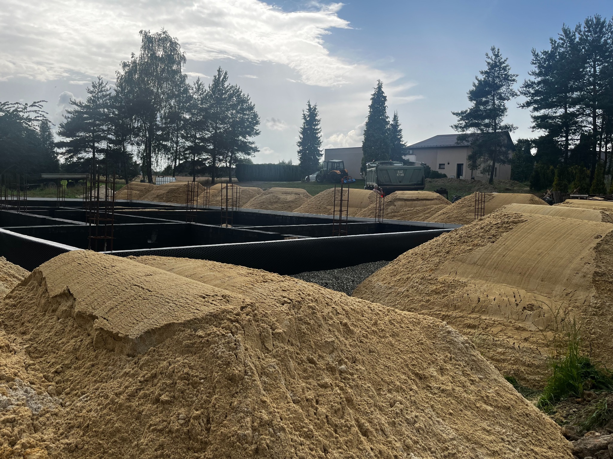 Przygotowanie terenu pod budowę: fundamenty zbrojone, hałdy podłoża, widoczny traktor i przyczepa w tle, słoneczny dzień.