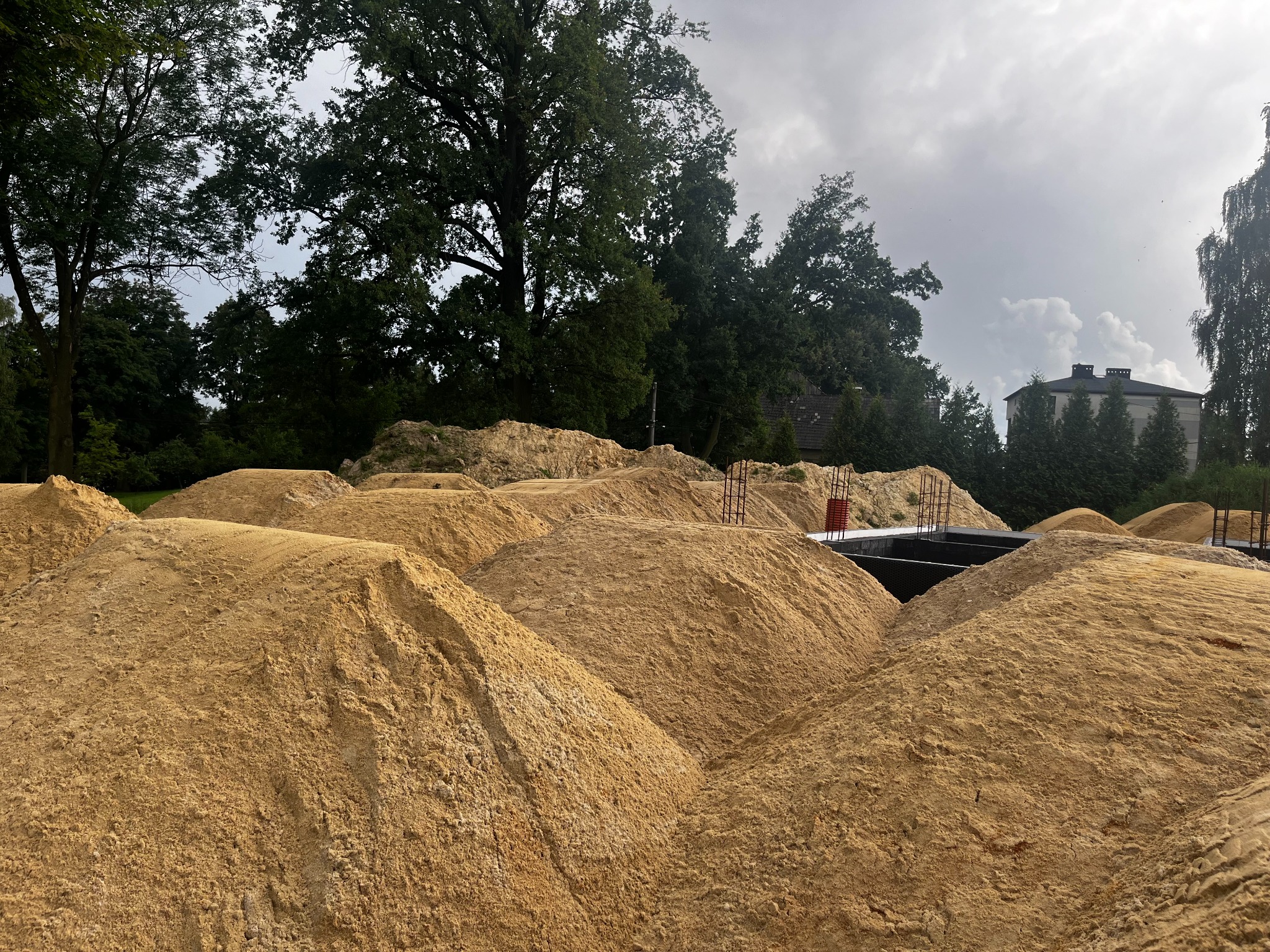 Spora ilość piasku budowlanego usypana w pryzmy na placu budowy z widocznym zbrojeniem i fragmentem fundamentów w tle, w otoczeniu zieleni.