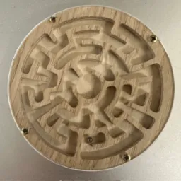 Drewniany labirynt z metalową kulką wewnątrz, wykonany z precyzją. Widoczne śruby mocujące. Jasne drewno i przezroczyste tworzywo.