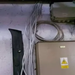 Białe przewody elektryczne prowadzone wzdłuż ściany, przymocowane do metalowej listwy. Widoczna skrzynka elektryczna z symbolem ostrzegawczym.