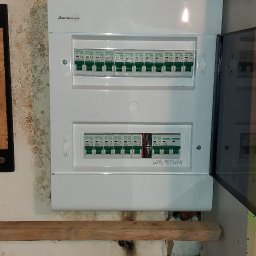 MD BUD - Rozdzielnica elektryczna marki Elektro-Plast Gacia z bezpiecznikami, częściowo otwarta, widoczne kable. Tło to ściana z widocznymi śladami wilgoci i prowizoryczna półka.