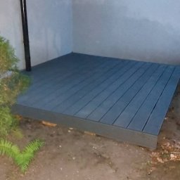 MD BUD - Szara, drewniana podłoga tarasowa z desek kompozytowych, ułożona przy ścianie budynku, widoczna z poziomu gruntu. Obok roślinność i rura spustowa.