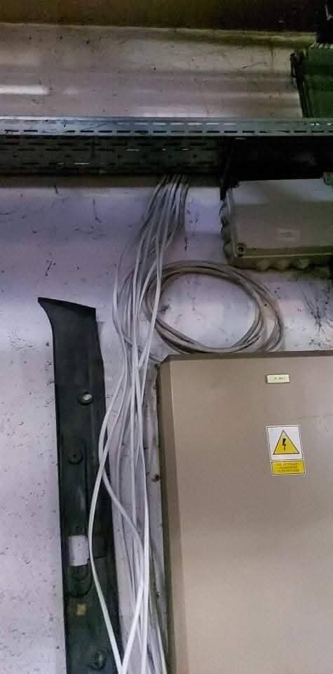 Białe przewody elektryczne prowadzone wzdłuż ściany, przymocowane do metalowej listwy. Widoczna skrzynka elektryczna z symbolem ostrzegawczym.