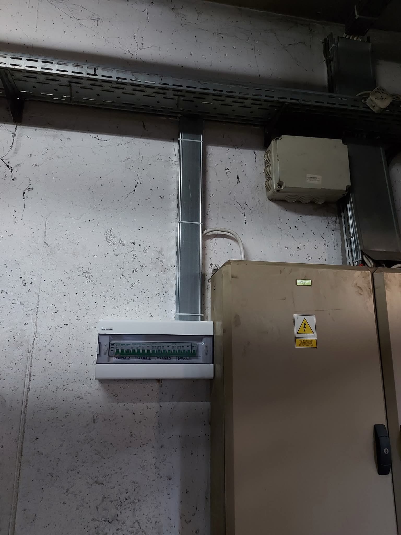 Szara skrzynka rozdzielcza z przewodami na ścianie w Łasku. Widoczna tabliczka ostrzegawcza i koryto kablowe.