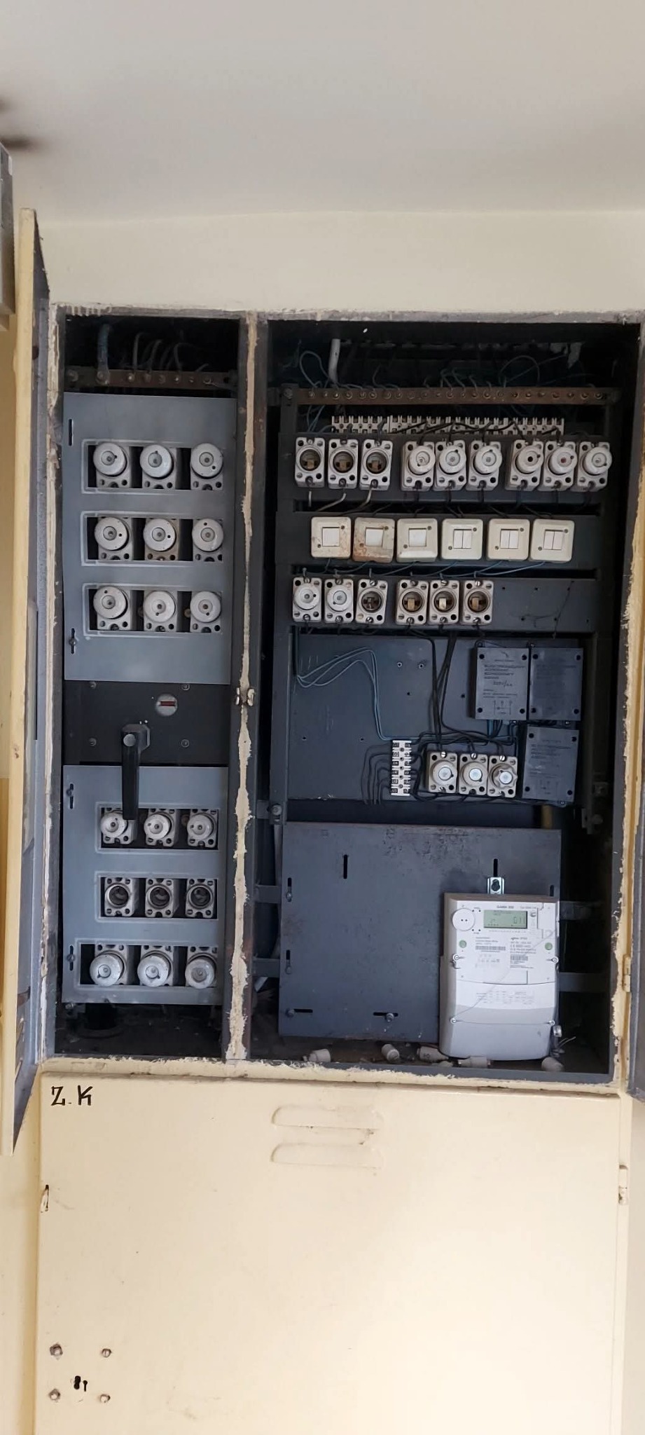 Otwarta skrzynka elektryczna z bezpiecznikami topikowymi i licznikiem energii elektrycznej wewnątrz, widoczne okablowanie i elementy instalacji.