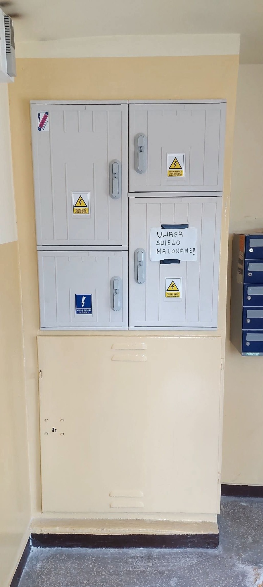 Szara skrzynka elektryczna z naklejkami ostrzegawczymi i kartką 'Uwaga! Świeżo malowane!' w korytarzu budynku. Widoczne skrzynki pocztowe.
