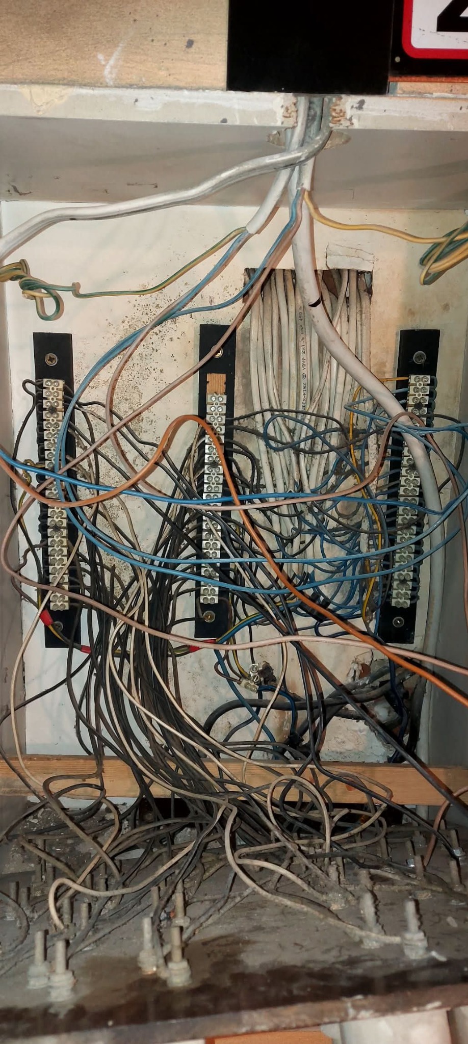 Otwarta skrzynka elektryczna z widocznymi kablami różnej grubości i koloru, podłączonymi do listew zaciskowych. Widoczne ślady zabrudzeń i kurzu.