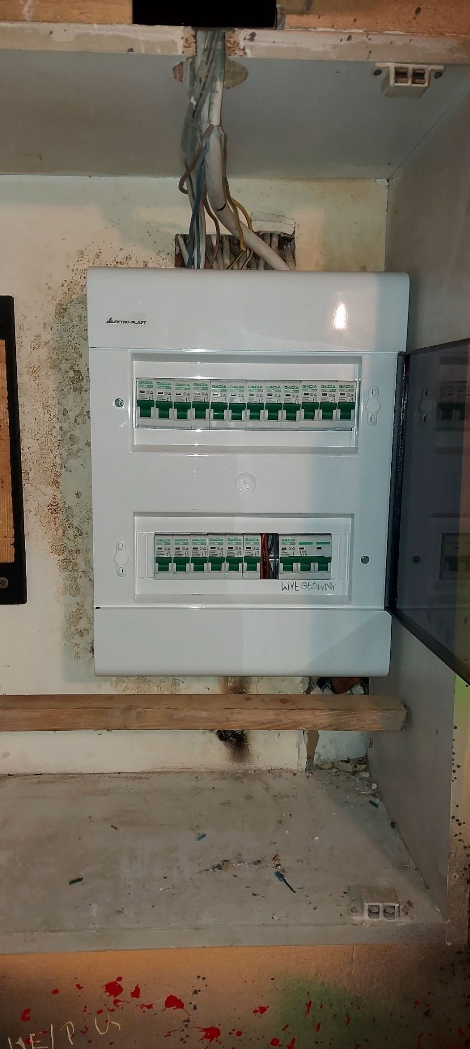 Rozdzielnica elektryczna marki Elektro-Plast Gacia z bezpiecznikami, częściowo otwarta, widoczne kable. Tło to ściana z widocznymi śladami wilgoci i prowizoryczna półka.