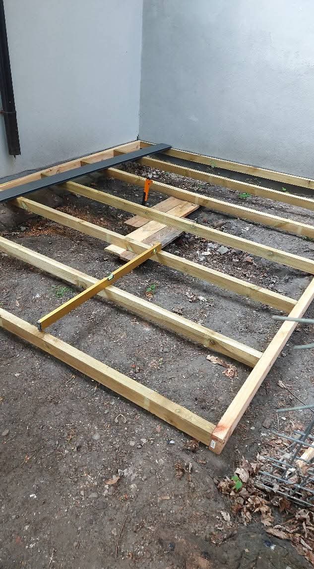 Drewniany szkielet pod taras na ziemi, w trakcie budowy, poziomowany poziomicą, z widocznym fragmentem ściany budynku. Elementy konstrukcyjne w surowym drewnie.