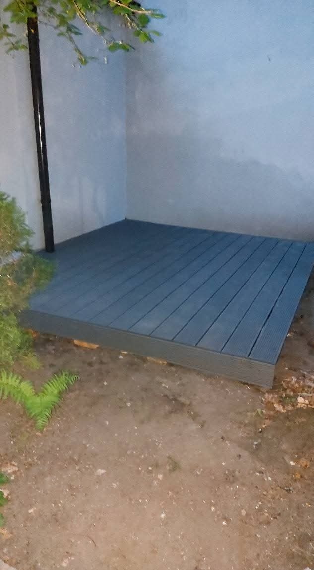 Szara, drewniana podłoga tarasowa z desek kompozytowych, ułożona przy ścianie budynku, widoczna z poziomu gruntu. Obok roślinność i rura spustowa.