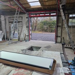 Bramy Ogrodzenia i Automatyka Paweł Chyrek - Montaż bramy garażowej: pracownik na drabinie instaluje prowadnice, w tle widoczna brama w opakowaniu, narzędzia i surowy garaż z otwartym wjazdem na zewnątrz.