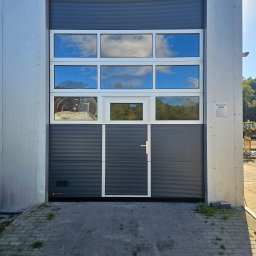 Bramy Ogrodzenia i Automatyka Paweł Chyrek - Szara brama segmentowa z oknami, w których odbija się niebo. Widoczny fragment wnętrza magazynu przez jedno z okien. Na ścianie tabliczka informacyjna.