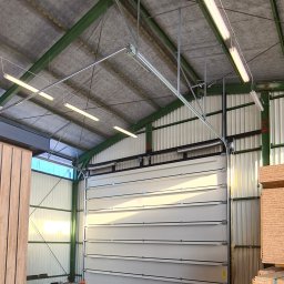 Bramy Ogrodzenia i Automatyka Paweł Chyrek - Zamknięta, segmentowa brama przemysłowa w hali z widoczną konstrukcją dachu i oświetleniem. Po bokach składowane płyty drewniane.