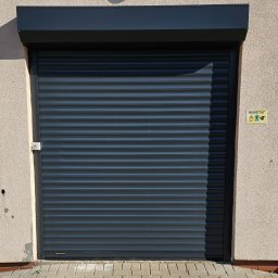Bramy Ogrodzenia i Automatyka Paweł Chyrek - Zamknięta, grafitowa brama rolowana z widocznym logo producenta u dołu, zamontowana w budynku warsztatowym z lampą nad bramą i kostką brukową u dołu.