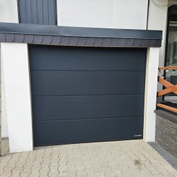 Bramy Ogrodzenia i Automatyka Paweł Chyrek - Zamknięta, segmentowa brama garażowa Hörmann w kolorze antracytowym, z poziomymi przetłoczeniami, zamontowana w białej wnęce budynku. Widoczny fragment podjazdu z kostki.