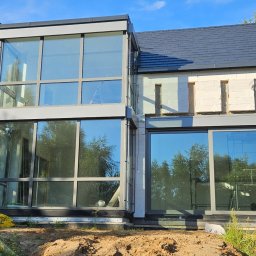 Bramy Ogrodzenia i Automatyka Paweł Chyrek - Dom w trakcie budowy z dużymi oknami i szarą stolarką. Widoczne niedokończone ściany z białego bloczka i przygotowany teren wokół budynku.