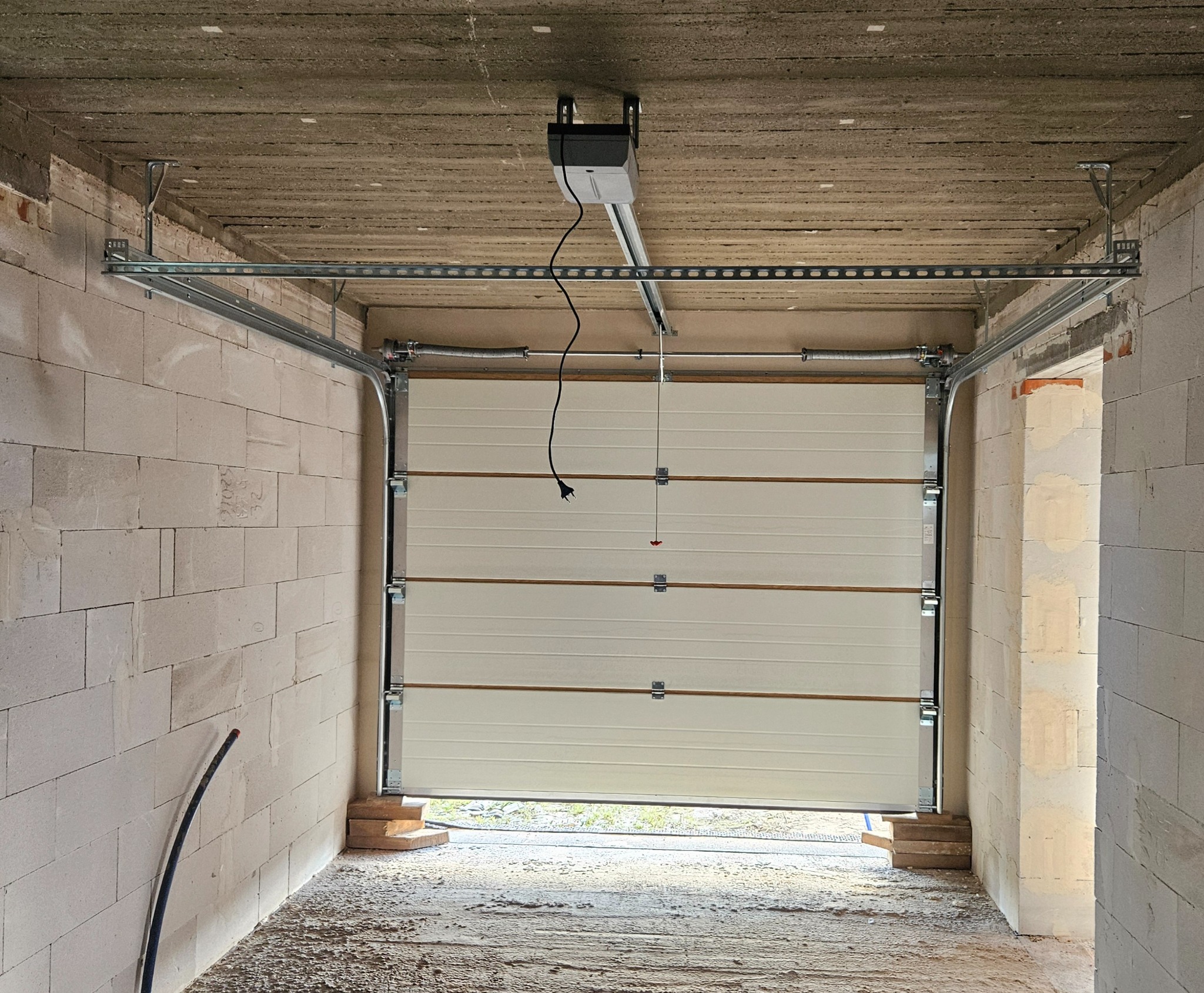 Biała segmentowa brama garażowa z automatyką w surowym garażu z betonu komórkowego. Widoczne prowadnice, sprężyny i silnik. Częściowo otwarta, z widokiem na zewnątrz.