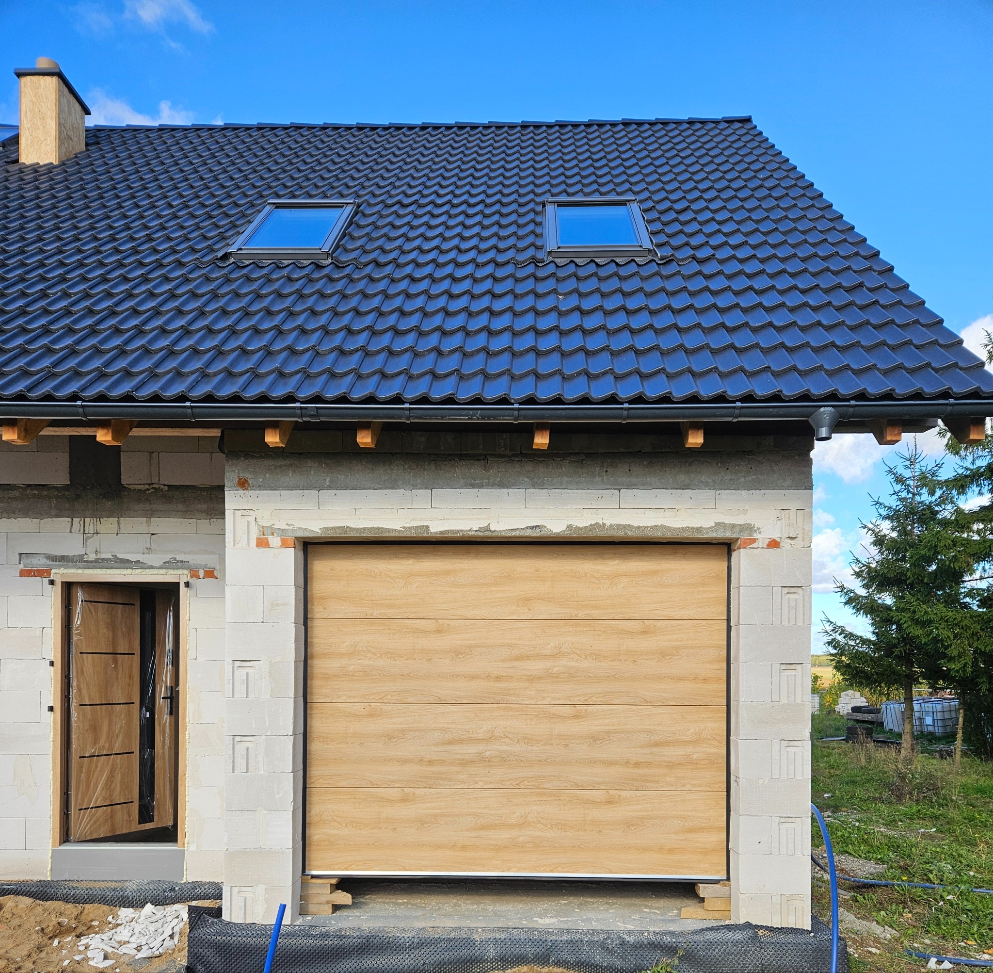 Jasna brama garażowa segmentowa z imitacją drewna w surowym budynku z białego bloczka, widoczne okna dachowe i częściowo otwarte drzwi wejściowe.