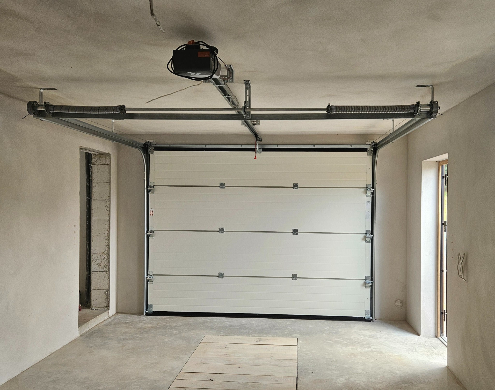 Biała segmentowa brama garażowa z widocznym mechanizmem sprężynowym i napędem, zamontowana w surowym garażu z betonową podłogą i oknem.