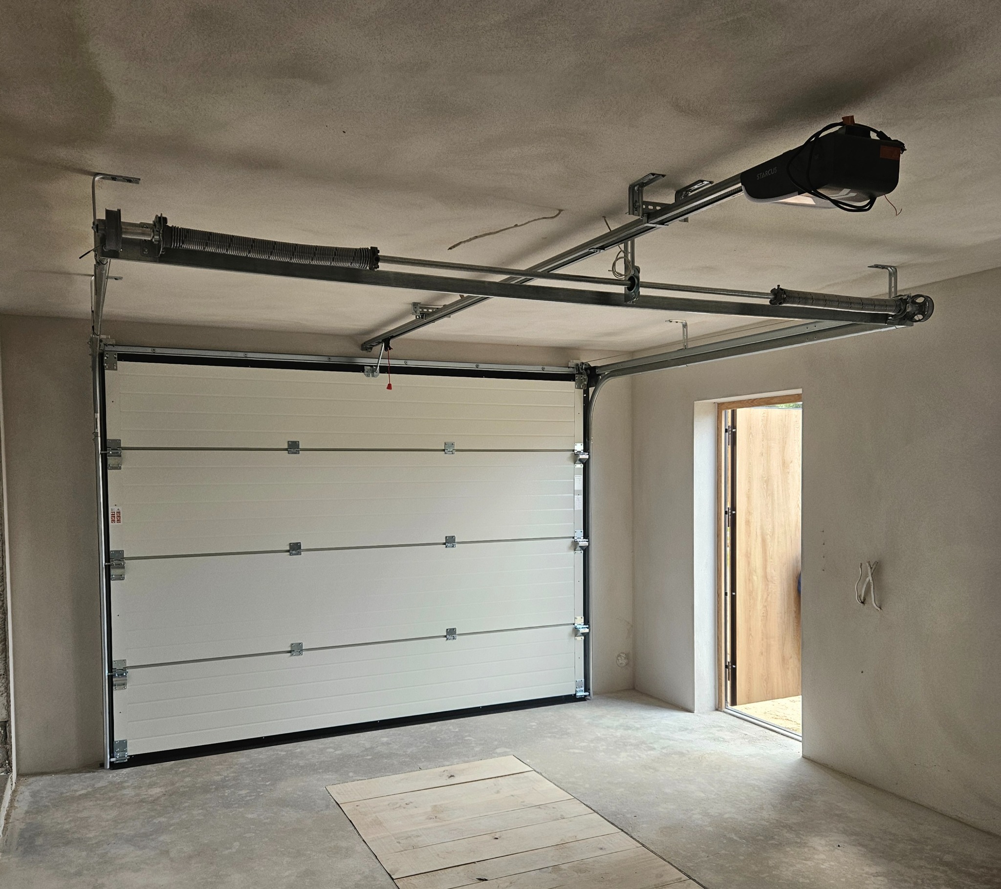 Zamknięta, segmentowa brama garażowa z napędem Starcus, widoczna od wewnątrz w surowym garażu w Słupsku. Białe panele bramy, szara konstrukcja nośna.