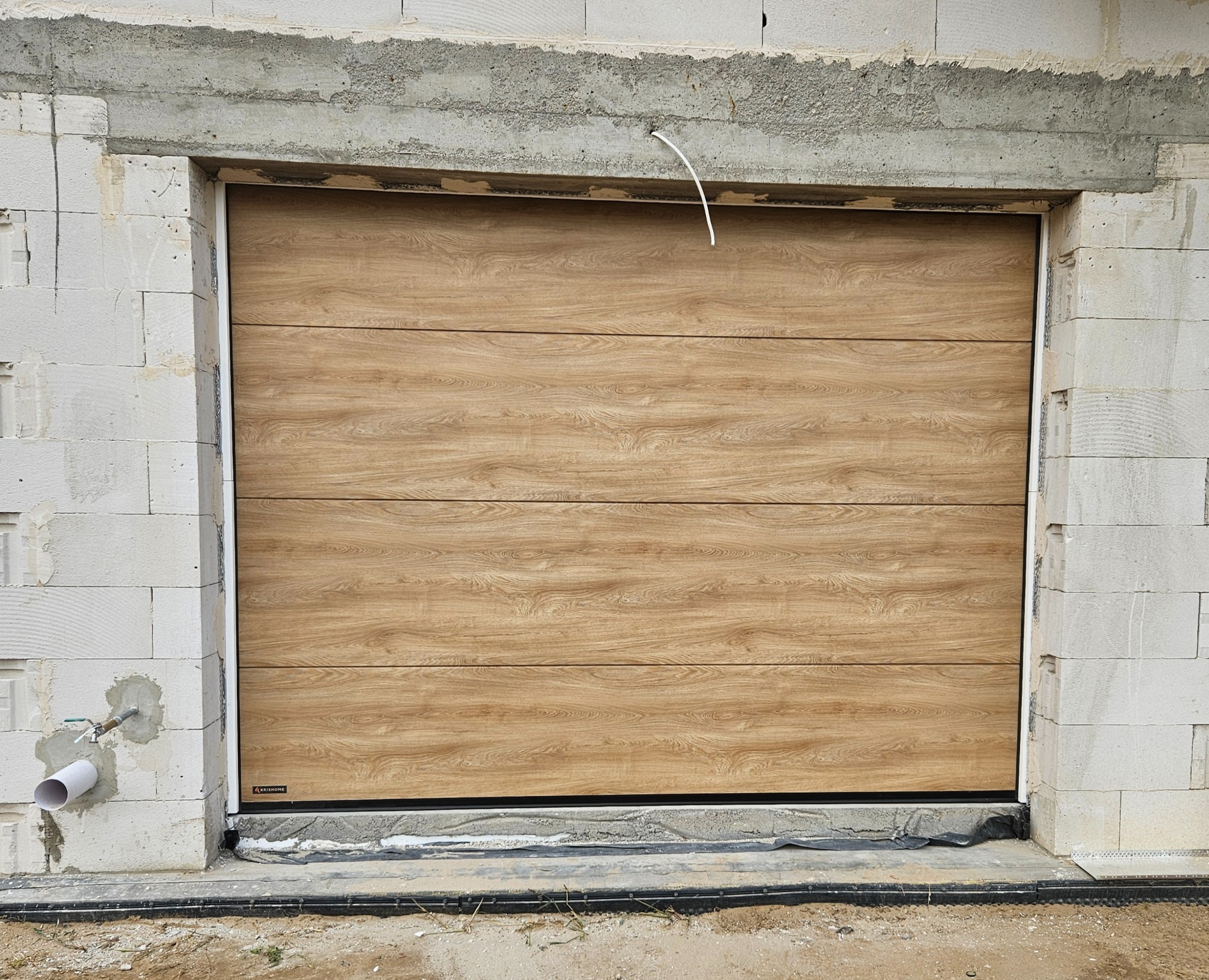 Zamknięta segmentowa brama garażowa z poziomym wzorem drewna, zamontowana w surowym otworze budynku z białego betonu komórkowego. Widoczny przewód instalacji elektrycznej.