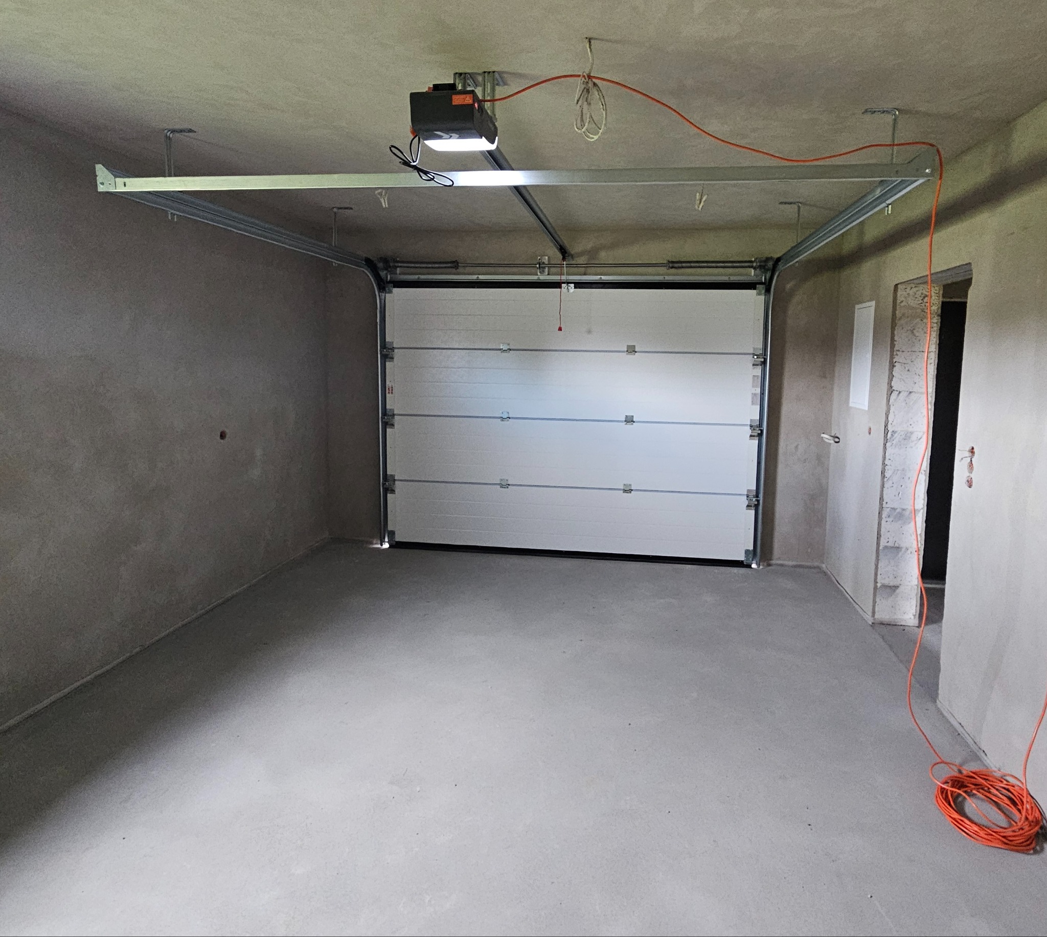 Wnętrze surowego garażu z zamontowaną białą segmentową bramą. Widoczne szyny, napęd z oświetleniem i zwisający pomarańczowy kabel. Ściany i podłoga w stanie deweloperskim.