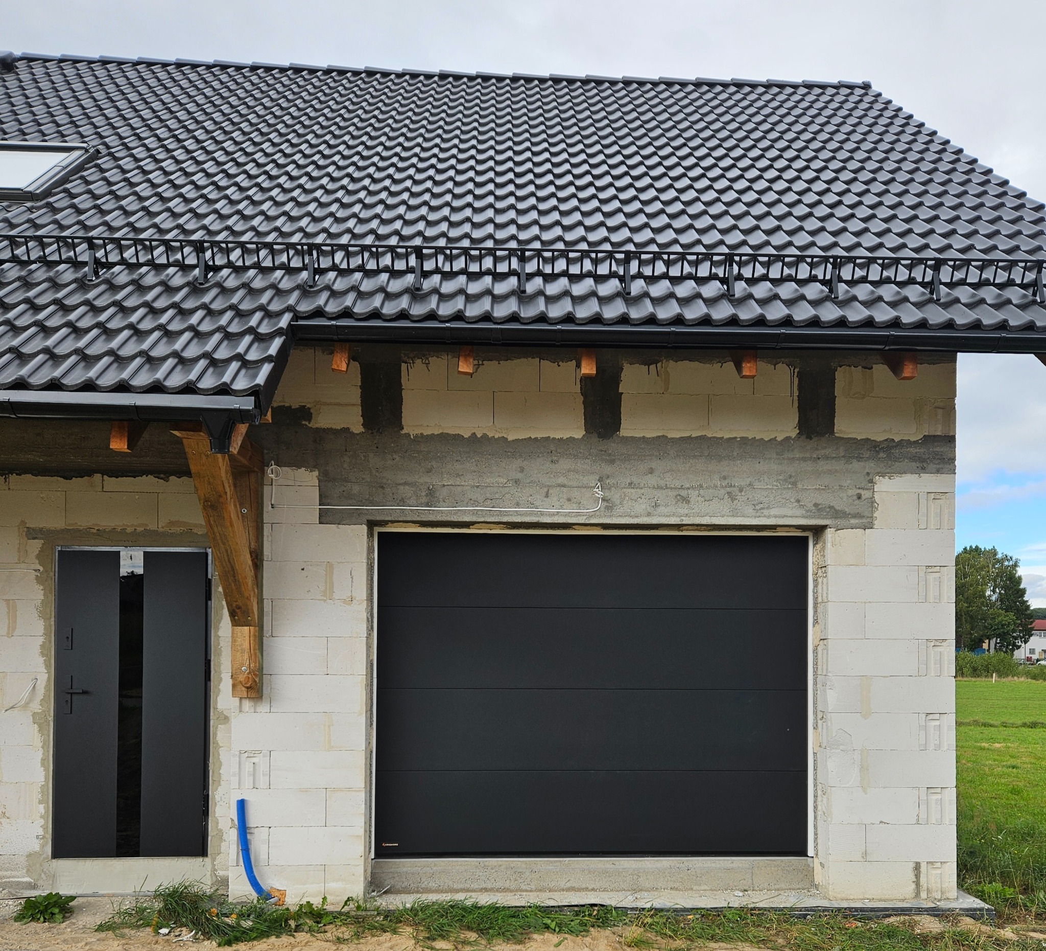 Czarna brama garażowa segmentowa w surowym otoczeniu budynku w trakcie budowy, widoczna brama wejściowa obok. Dach pokryty czarną dachówką.
