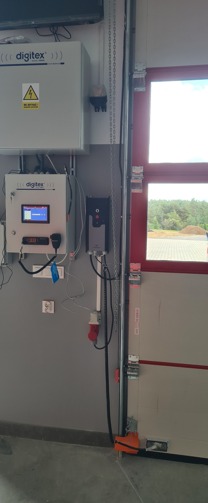 Panel sterowania bramą garażową z wyświetlaczem i radiotelefonem, zamontowany na szarej ścianie obok czerwonej ramy bramy. Widoczne okablowanie i elementy automatyki.