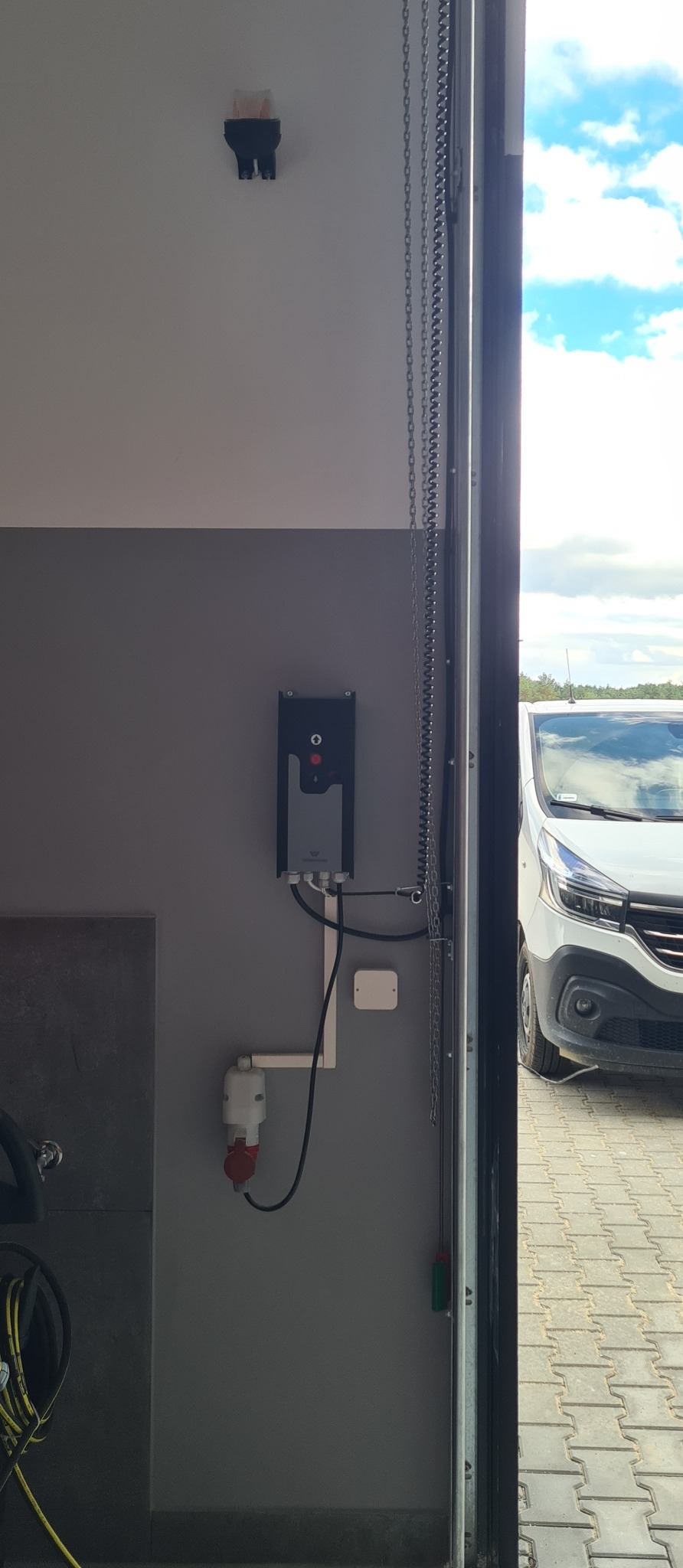 Panel sterowania automatyką bramy garażowej na szarej ścianie, widoczny fragment łańcucha i brama otwarta na zewnątrz, gdzie stoi biały samochód dostawczy.