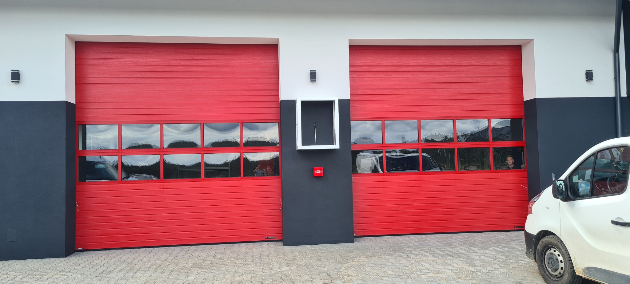 Dwie czerwone bramy garażowe segmentowe z oknami, oddzielone szarą kolumną z białą ramką i czerwonym przyciskiem. Biały samochód częściowo widoczny po prawej stronie.
