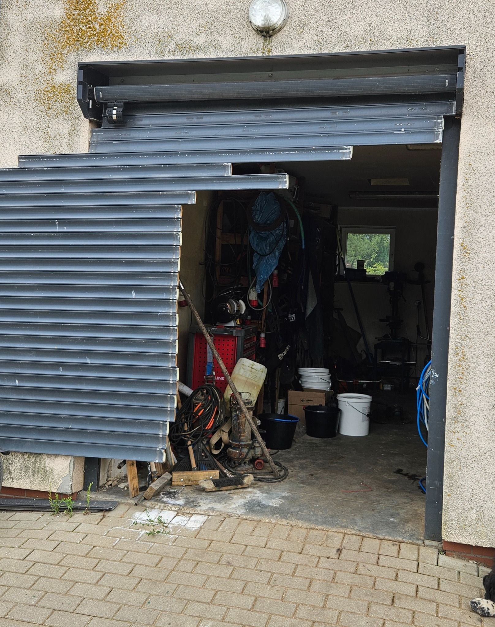 Uszkodzona brama garażowa z blachy falistej, częściowo otwarta, ukazująca wnętrze zagraconego garażu. Widoczne uszkodzenia mechaniczne i brakujące elementy.