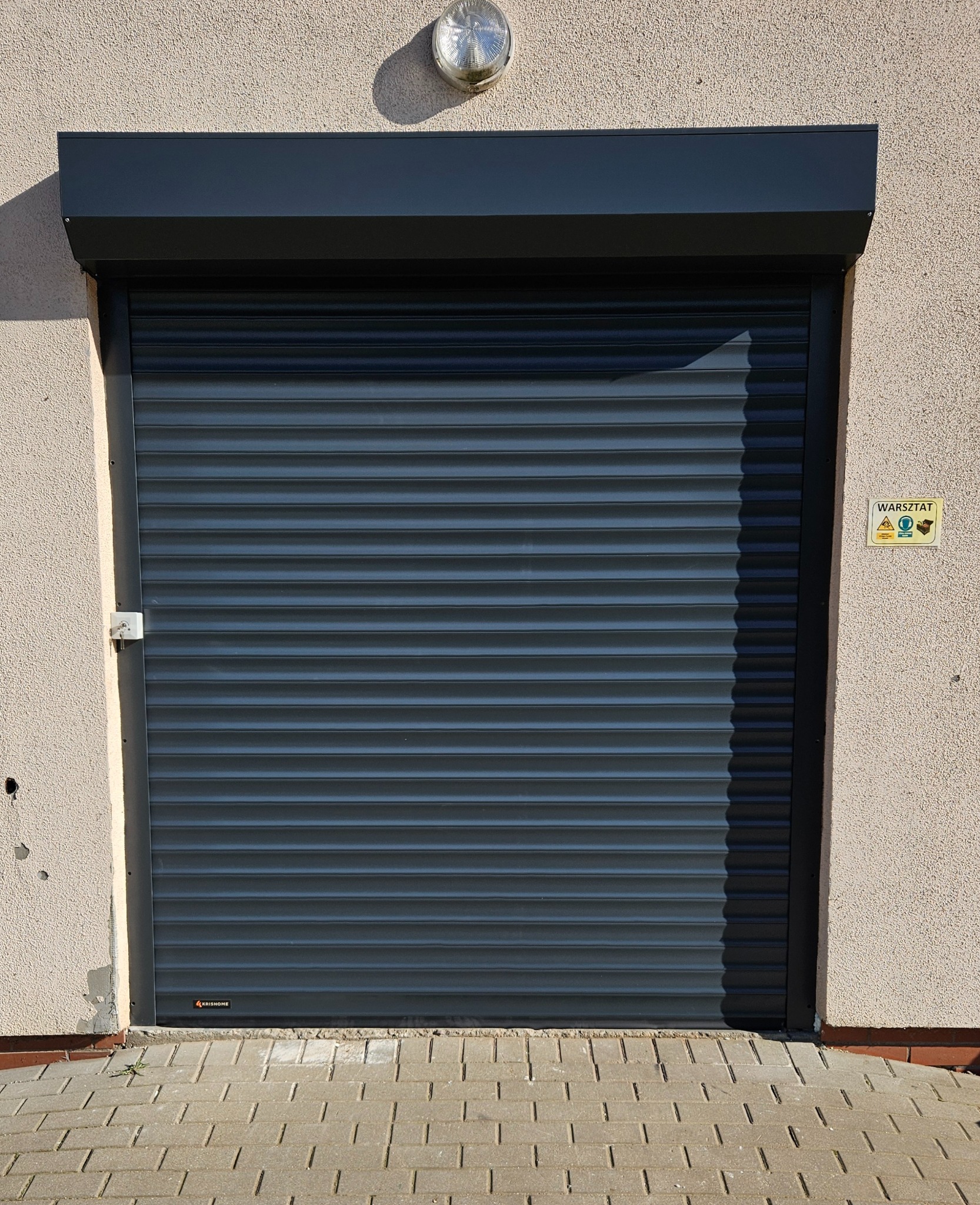 Zamknięta, grafitowa brama rolowana z widocznym logo producenta u dołu, zamontowana w budynku warsztatowym z lampą nad bramą i kostką brukową u dołu.