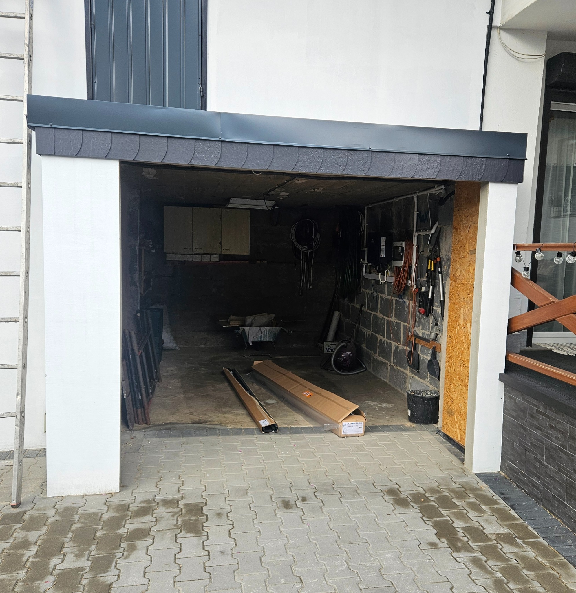 Otwarty garaż z widocznymi elementami w trakcie montażu. Wnętrze garażu z narzędziami i materiałami budowlanymi. Widoczna drabina oparta o ścianę.