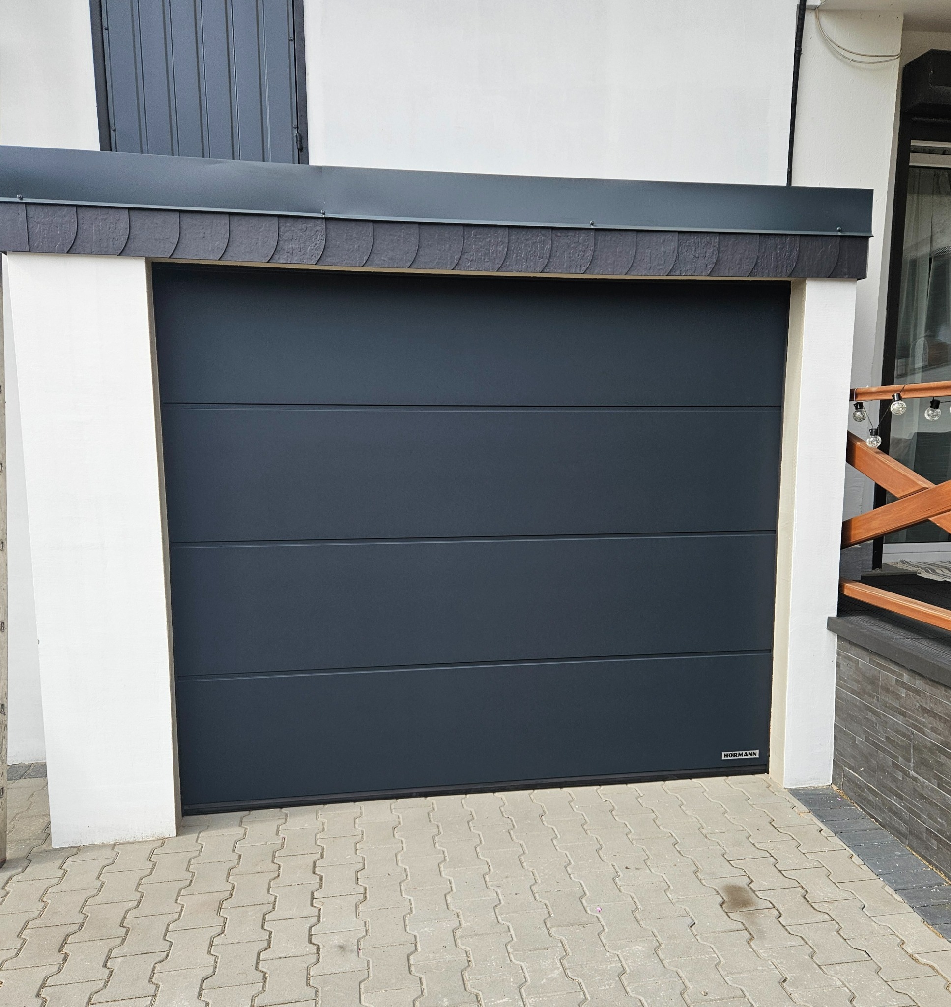 Zamknięta, segmentowa brama garażowa Hörmann w kolorze antracytowym, z poziomymi przetłoczeniami, zamontowana w białej wnęce budynku. Widoczny fragment podjazdu z kostki.