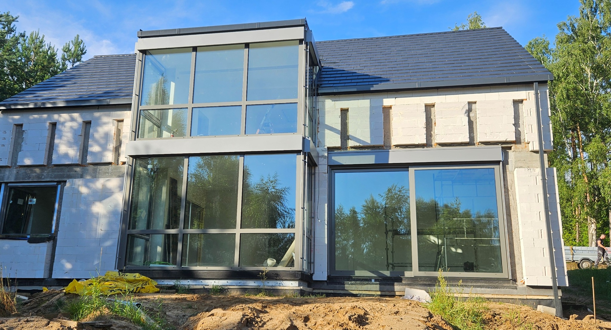 Dom w trakcie budowy z dużymi oknami i szarą stolarką. Widoczne niedokończone ściany z białego bloczka i przygotowany teren wokół budynku.